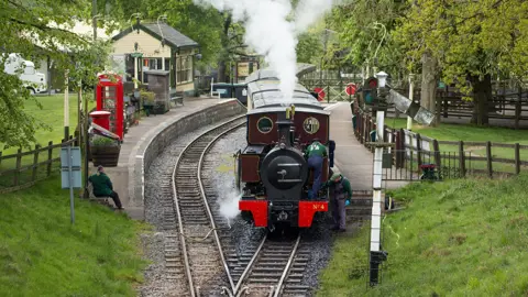 An historic steam train waiting at the platform at Whipsnade Zoo railway. It is surrounded by mature trees and grassy embankments.