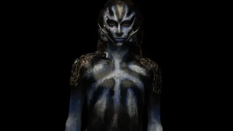 Model with black and blue make-up on their face and body, with prosthetic ears attached to their face.