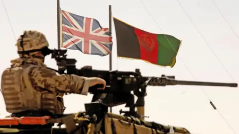 Getty Images A soldier on a vehicle with a mounted gun. The flags of the United Kingdom and Afghanistan are flapping in the wind. 