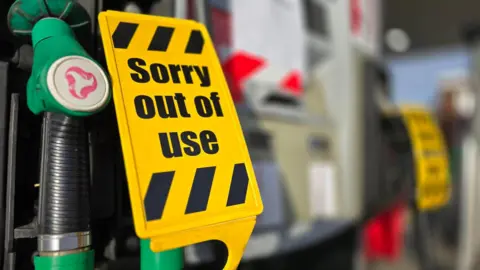 A yellow "sorry out of use" sign over green unleaded petrol pump nozzles.