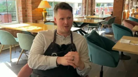 A man sits in a chair in a restaurant with his hands clasped together. He is wearing a dark apron over a lighter coloured shirt