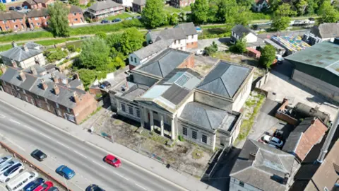 National Lottery Heritage Fund Aerial shot of an old building with pillars at the front and houses at either side. A road with passing cars is in front.
