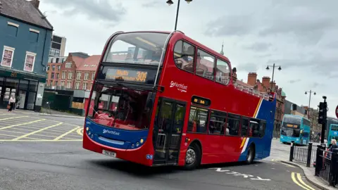 A red and blue, double-decker open top bus driving on a road. There are two blue buses behind it. The front of the bus reads 'Blyth 309' and has the Go North East logo below the windscreen. There are buildings to the left behind the bus.