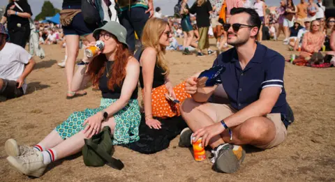 PA Media People enjoy the warm weather at the Forwards Festival at the Downs in Bristol. Two women and a man sit on the ground drinking from tins and fanning themselves.