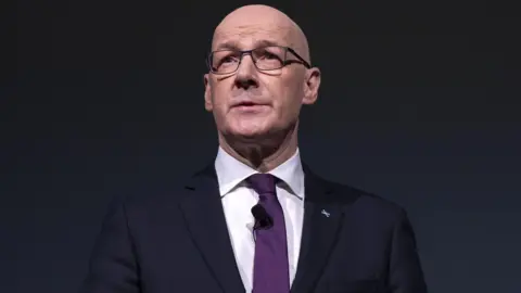John Swinney wearing a dark suit over a white shirt and purple tie standing against a black background with his hands interlocked in front of him.