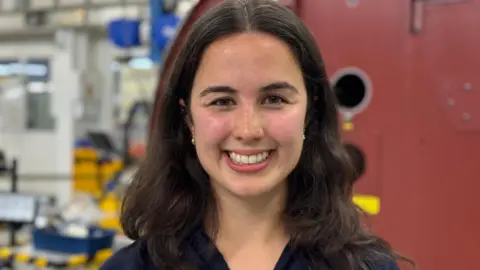 Lucinda Adam/BBC Ella has long wavy dark brown hair and dark brown eyes. She wears a navy blue polo shirt. She stands on the factory floor of Elekta, a medical equipment manufacturer.