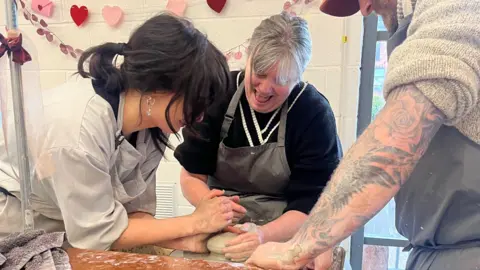DAIZY STEVENS Two women and one man stand around a potters wheel, all with their hands in the clay. The woman on the left has black hair, a white shirt with rolled up sleeves and a white apron. The woman in the middle is laughing and has grey hair, a grey apron and a black jumper with rolled up sleeves. The man on the right has a tattooed arm, woolly jumper with rolled up sleeves and a grey apron. They are throwing the pot in a white studio with windows.