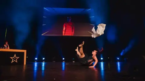 Homecare's Got Talent A woman sitting on a stage with her legs in the air and hands on the floor as a white piece of cloth flies above her head. A judge can be seen sitting at a table on the left and a large screen overlooks the stage.