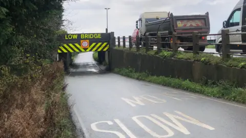 John Devine/BBC Stonea Road bridge near Manea. It shows two parallel roads, one going under the railway line with slow written in yellow writing on the tarmac. The bridge has yellow signs saying low bridge and yellow warning stripes. Beside it and rising above to the railway line, a flat-bed lorry and a van can be seen waiting to cross the railway line once the barrier is lifted