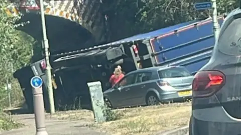 Supplied A large blue lorry that has overturned in front of a railway bridge. Surrounding cars are stationary.