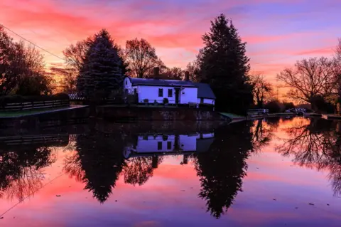 BBC Weather Watchers/No1GhostDog A sunrise of dappled pinks and purples frames a white waterside building next to a canal basin. The scene is perfectly reflected in the still water. 