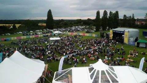 Venue 16/Summer Soul Vibes A drone image of Summer Soul Vibes. Attendees are pictured in a large field with a stage at one end. Other tents can be seen dotted around the outside of the venue.