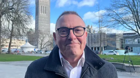 Plymouth City Council leader Tudor Evans smiles while standing outside the Civic Centre. Plymouth's Crown Court and Guildhall can be seen in the background.