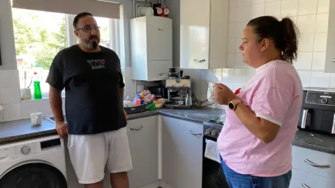 A man and woman stood facing each other in the kitchen of their home. The man is stood by the washing machine wearing a black logo t-shirt and light grey shorts. The woman is stood opposite him wearing a pink t-shirt and blue jeans. She is holding a mug. 