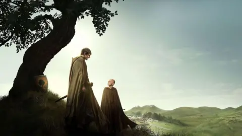 Publicity shot from A Knight of the Seven Kingdoms. Shows Ser Duncan the Tall and Egg standing beside a curved tree with mountains in the background. Ser Duncan is wearing a light coloured cloak, has dark brown hair and carrying a sword looking down at Egg. Egg is a young child and is bald, wearing a light coloured cloak and looking up at Ser Duncan.