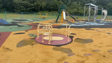 BBC/Claire Hamilton A dilapidated children's playground with a red and yellow roundabout in the foreground, and grey slides and climbing frames in the background. 