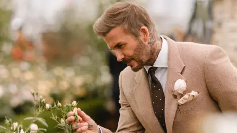 Courtney Louise Photography A man with short blond hair and a beige suit leaning over a bush and examining a flower