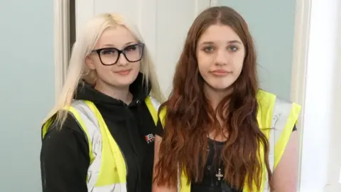 Two teenagers in dark tops and hi-vis vests stand against a light blue wall. On the let, Victoria has long blonde and and glasses. On the right Karys has long reddish brown hair.