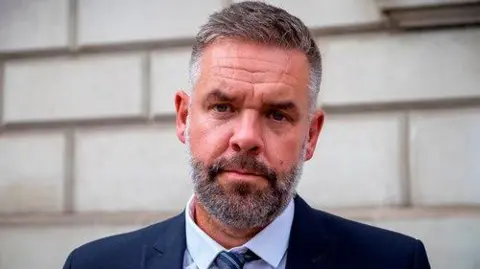 Humberside Police Federation Lee Sims, the chair of the Humberside Police Federation, stands in front of a stone brick wall. He is wearing a dark blue suit, blue shirt and a grey and blue striped tie. He has short cropped, greying hair and a beard.