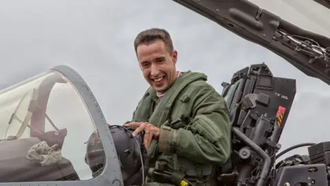 LONG FAMILY A pilot in a green flying suits smiles as he sits in the cockpit of an RAF jet, with the canopy raised above his head.