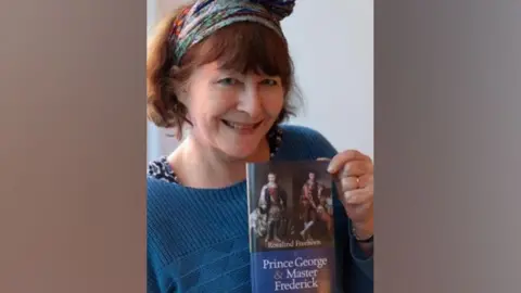 Rosalind Freeborn is wearing a blue jumper and is smiling at the camera. She is holding a copy of her book, Prince George and Master Frederick.