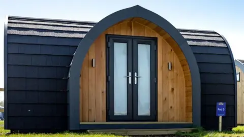 A camping pod in a holiday park field. It had black tiles on its exterior and wooden panelling in its porch area along with two glass doors with black trim.