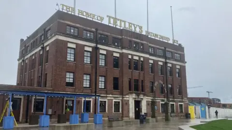 A large five-floor red brick building with gold lettering on the roof that reads 'The Home of Tetley's Established 1822'. It is a grey day.