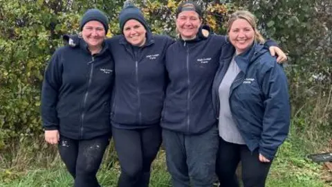 High Lodge Farm Katy, Jess, Lucy and Emily stand side by side with their arms around each other, facing into the camera with big smiles on their faces. All four are wearing navy fleeces and waterproof jackets bearing a small High Lodge Farm logo in white.