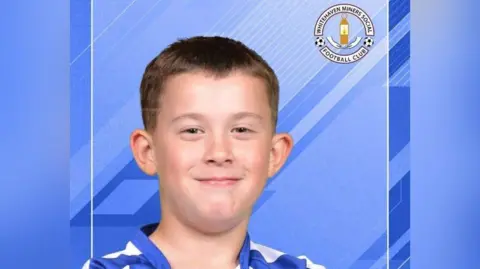 Apostila para a família Jay Cartmell, com uma camisa de futebol azul e branca, olhando para a câmera e sorrindo. Atrás dele está um fundo azul e um emblema do Whitehaven Miners Football Club no canto superior direito.