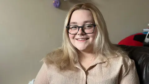 A woman with blonde hair and black glasses smiles at the camera. She has a nose piercing and an eyebrow piercing, and wears a beige button-up shirt