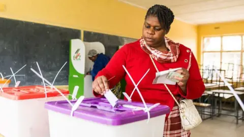 Inside a school classroom in Lusaka, Zambia, a woman wearing a red top places a ballot paper in a plastic ballot box with a purple lid. Some desks can be seen behind her. 