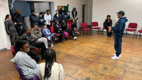 Children and teenagers are sitting in a semi-circle as Adam Deacon gives acting tips