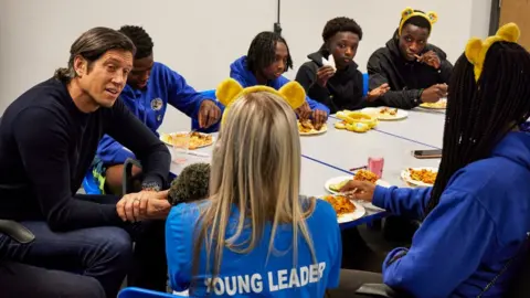 Vernon Kay and six others sitting around a table. He is interviewing one of them, a woman with a T-shirt saying "young leader".