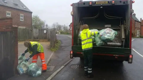 A man with a hi-vis vest picking up rubbish bags and another man with a hi-vis vest putting bags into a bin lorry. It is a residential street with houses.