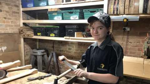 BBC/Alan Webber Sophie smiling into the camera as she works on a piece of wood in a workshop wearing an Adidas cap and Garners polo-shirt with a ponytail