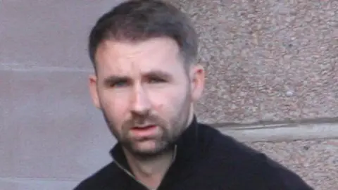 Tim Bugler James Keatings looking straight at the camera. He is wearing a black zip neck jumper. He has short, dark hair and a short, dark beard.