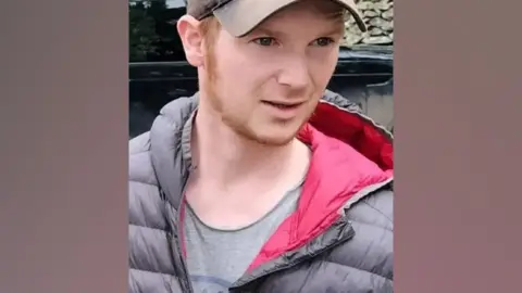 Thomas Scaife. He has a thin ginger beard and is wearing a baseball cap and grey jacket with red lining. He is looking to his right.