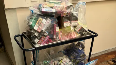 Multiple tobacco and vape products in clear plastic bags on a black trolley. 