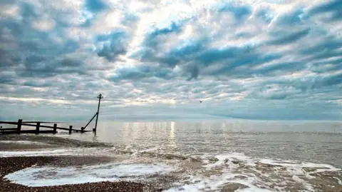 A view of the coast at Felixstowe on a cloudy day. Some sun can be seen peaking through the clouds above the sea. 