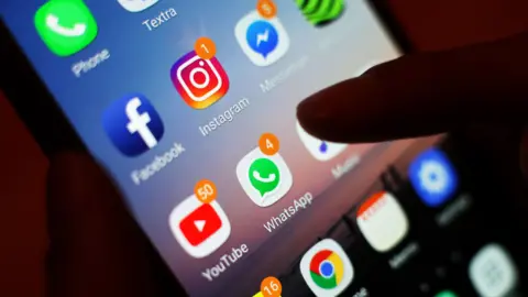 Stock image of a thumb scrolling through a mobile phone. There are several icons on the smart phone, including Whatsapp, YouTube and Instagram.