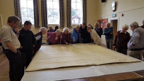 About 11 people gather around the tithe map some pointing at it as it is on display on a large table in a village hall 