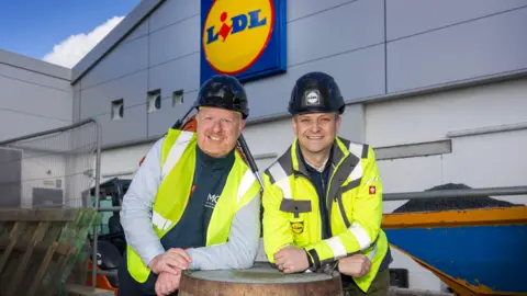 Lidl Two men with black hard hats, high vis jackets leaning on a barrell. The Lidl logo is behind them