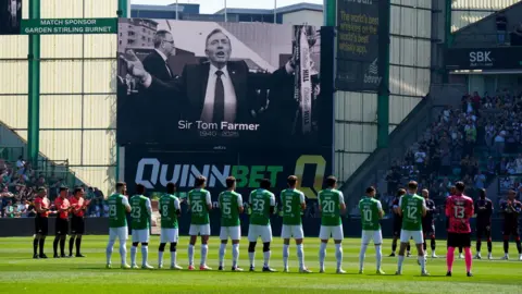 PA Media Footballers from Hibernian FC stand in a line clapping during a minute's applause with a picture of Sir Tom Farmer on a big screen behind him