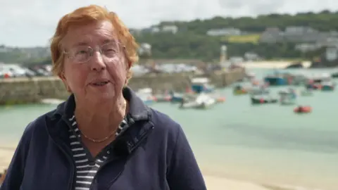 The image shows a person standing on a beach in front of a harbour filled with small boats, with a stone harbour wall and green hills dotted with houses in the background. The person is wearing glasses and a dark jacket, facing the camera, while the water behind is calm under a cloudy sky.