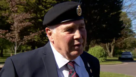 A man in a black beret, navy blazer, white shirt and red and black stripy tie is looking to the side with a serious expression. Behind him appears to be a car park with one white car parked, surrounded by greenery and trees.
