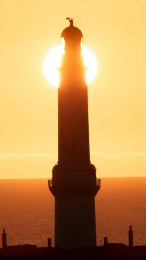 Lee Shirreffs A lighthouse in silhouette stands directly in front of the rising or setting sun, creating a glowing halo around its tower above the sea.