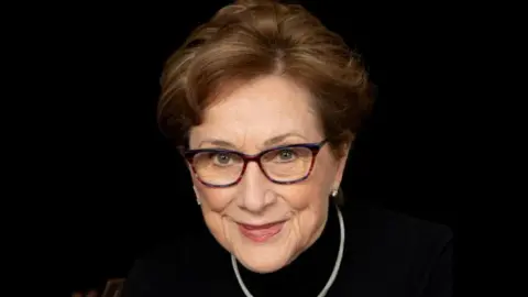 Richard Bailey A head and shoulders image of Professor Dame Carol Black, a woman with short, wavy brown hair and dark-rimmed glasses. She wears a silver necklace which contrasts with the black top she is wearing, against a black background.