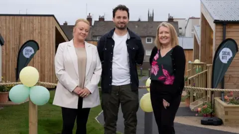Frame PR Three people - two women flanking a man - standing outside, in a garden with wood panelled buildings behind them. The first woman has blonde hair pulled back, a beige top and white jacket on. The man has a blue denim jacket and white shirt, with dark hair and a beard. The second woman has shoulder length blonde hair and a dark top with a pink and purple floral pattern across it.