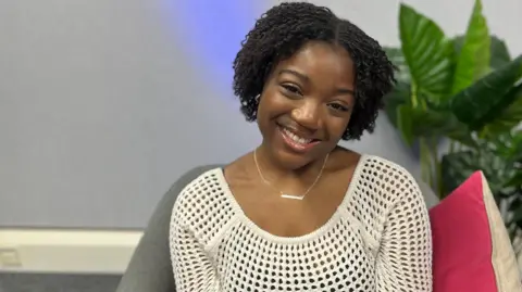 Lily is sat down and smiling. She is wearing a cream coloured top.