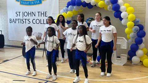 A group of girls doing a dance in a school gymnasium. A balloon arch is behind them.